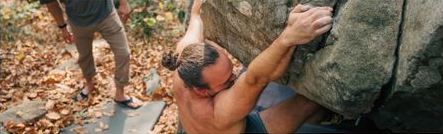An overhead angle of a shirtless man climbing a boulder outdoor.
